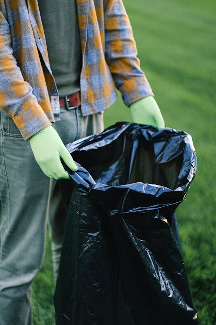 A person standing outdoors on a grassy area is holding open a large black rubbish bag, which appears to be made of plastic and lined with a shiny interior. The individual is wearing light green gloves