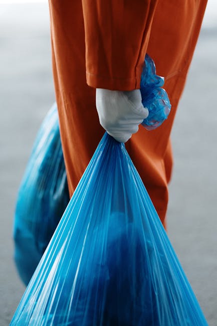 A close-up image of multiple black plastic rubbish bags, tightly sealed at the top with visible pleats and creases in the glossy material. The bags appear to be filled with household waste, placed on 