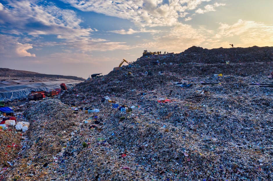 A large landfill site filled with a densely packed mound of mixed waste materials, including plastics, cardboard, textiles, and other refuse, covering a wide area and extending into the distance. The
